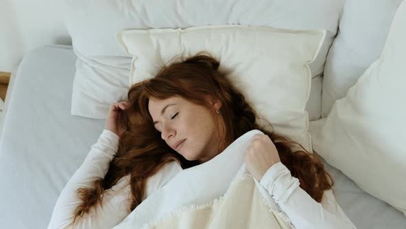 Calm attractive woman with red hair lies on a bed on a white pillow and sleeps