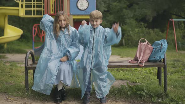 Cute Children Sitting on Bench on Playground and Putting on Raincoat Hoods alt