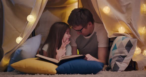 Happy Father and Daughter Lying on Floor in Teepee and Reading Book Together at Home alt