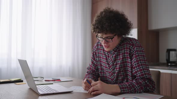Curly Haired Male Student Attractive Young Boy in Glasses is Studying at Home Using Laptop Typing alt