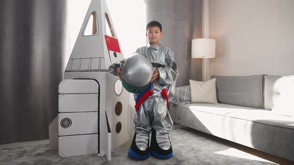 An Asian Boy in an Astronaut Costume Stands Near a Cardboard Model of a Spaceship and Puts on Helmet alt