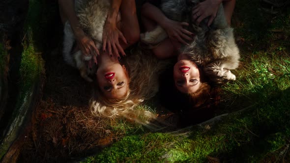 Two Women in Clothes Made of Natural Fur Lie on Grass in Forest and Sing a Song