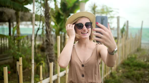 Cheerful Beautiful Girl in a Summer Hat and Glasses Calls for a Video Call on the Background of the