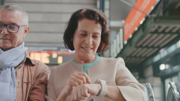 Cheerful Elderly Couple Chatting with Family on Mobile Phone and Smartwatch Waiting for Train alt