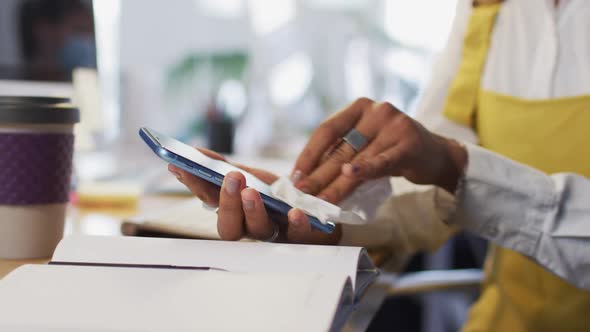 Woman wearing face mask wiping her smartphone with a tissue alt
