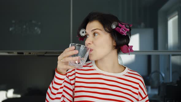 Woman with Hair Curlers Drinking Water in Kitchen alt
