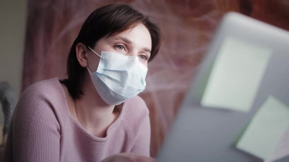 Young Woman in Medical Mask Studying or Working at Home Using Laptop Typing Message Writing Post alt