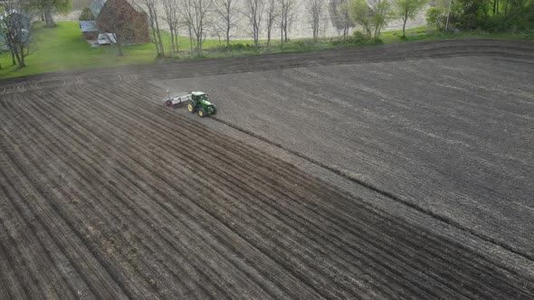 Farmer working field fertilizing and seeding with a dust cloud following. Barn behind row of trees.