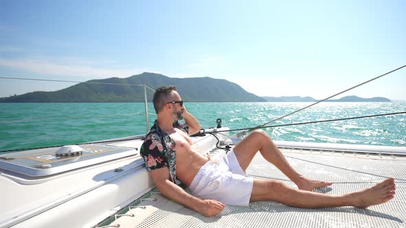 4K Caucasian man talking on phone for corporate business while catamaran sailing on summer vacation.