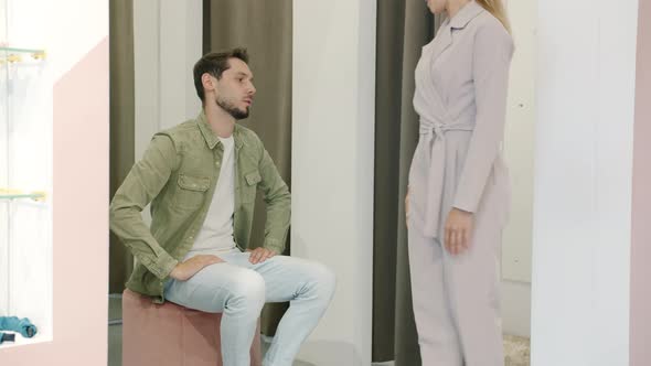 Annoyed Man Talking to Girlfriend Trying on Clothes in Fitting Room Not Happy with Shopping alt