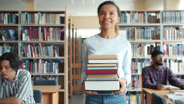 Pretty Mixed Race Lady Student Carrying Books Walking in Library Smiling alt