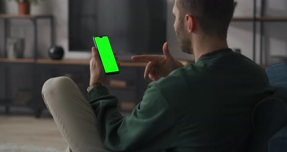 Man Is Chatting Online By Video Chat in Mobile Phone Sitting at Home on Couch in Living Room Green alt