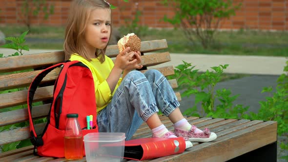 Girl Sits in Schoolyard and Eats Hamburger