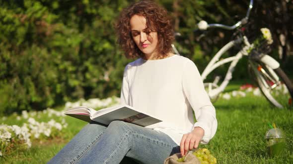 Young Woman in White Shirt and Blue Jeans is Reading a Book Sitting on the Grass in Park and Eating alt
