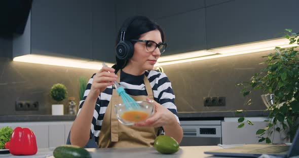 Woman in Earphones Enjoying Music while Whipping Eggs in Bowl Using whisk at the Table  alt