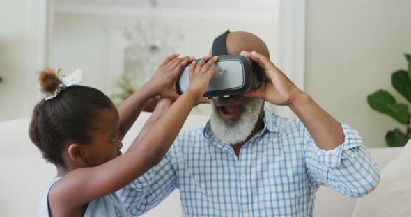 Happy african american grandfather using vr headset with granddaughter in living room alt
