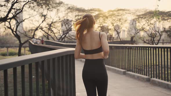 Woman in sportswear wearing a protective mask jogging exercise on the bridge in the city at sunset.