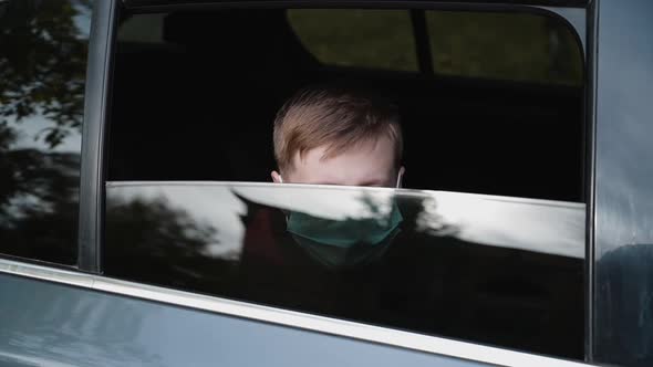 Young Boy in a Medical Mask Lowers the Glass and Looks Through the Car Window.