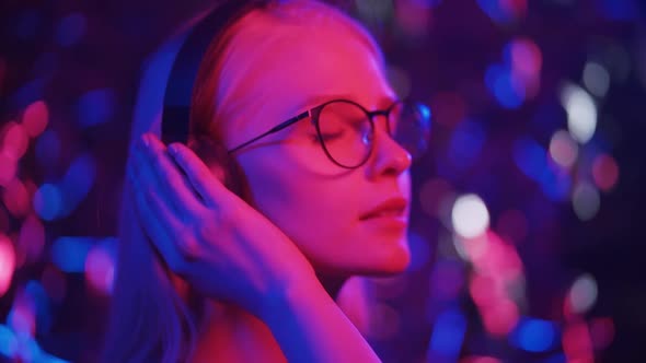 Happy Young Blonde Woman in Glasses Dances in Headphones on a Background of Multicolored Lights alt