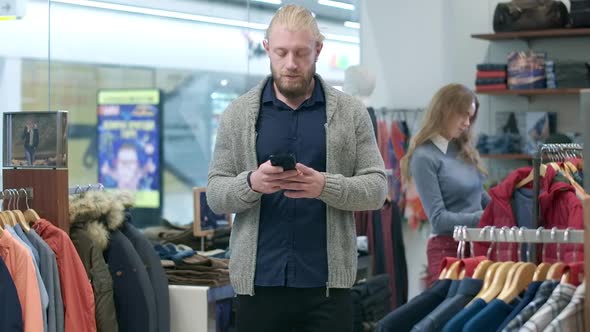 Handsome Young Bearded Man Surfing Internet on Smartphone While Woman Choosing Outfit at the alt