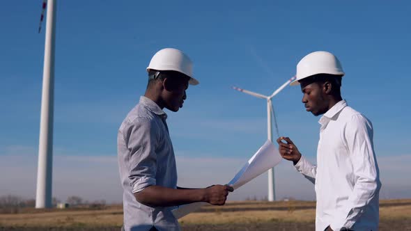 Two Male African American Electrical Engineers Stand Against the Backdrop of a Windmill at an Air alt
