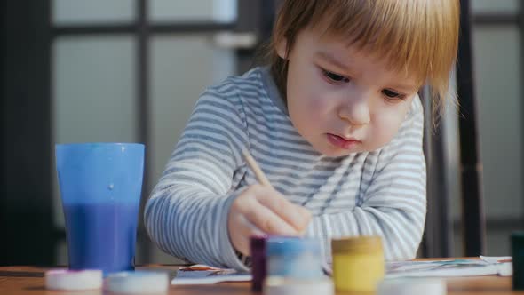A Cute Baby Boy is Engaged in Drawing with a Brush and Gouache Drawing with Paints on a Piece of