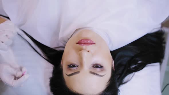 beautiful woman with black hair is lying on a couch on the procedure of permanent makeup lips alt