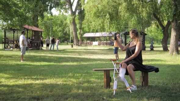 A Young Girl with a Cast and Crutches is Sitting in the Park Watching Friends Having Fun and Playing
