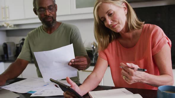 Diverse senior couple using tablet computer paying bills in kitchen alt