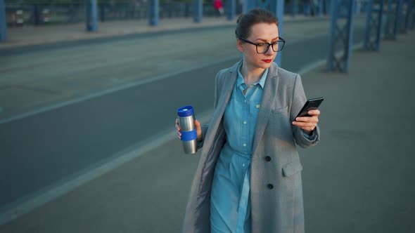 Woman in a Coat Walking Around the City in the Early Morning Drinking Coffee and Using Smartphone alt
