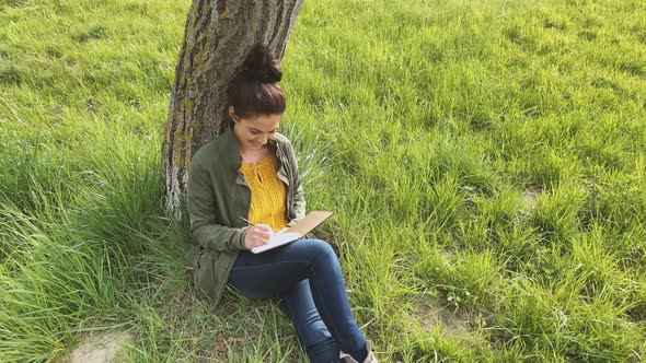 Young woman writing in notebook, sitting on meadow