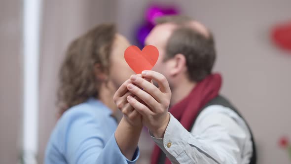 Closeup of Red Paper Heart in Male and Female Hands with Blurred Adult Couple Kissing at Background alt