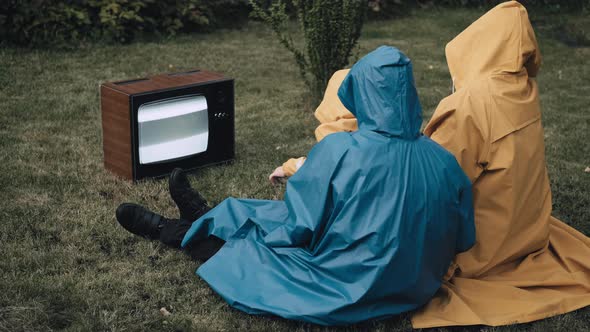 Family of Three is Sitting on the Grass in Raincoats and Watching Old Retro TV