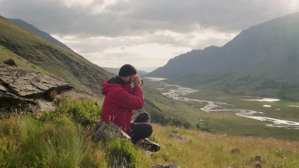 A young adventure photographer takes pictures around. Sitting on a mountain, against the backdrop alt