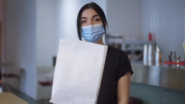 Medium Shot of Young Confident Woman in Covid Face Mask Stretching Packed Lunch to Camera Standing alt