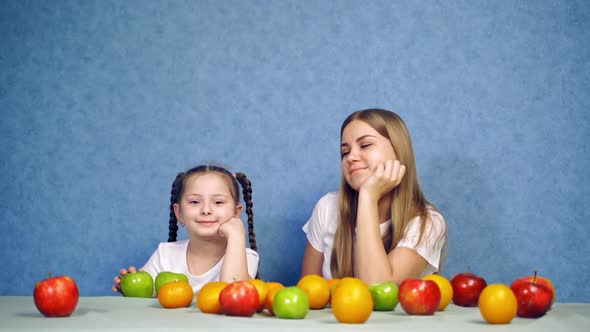 Girls sitting at table with fruits. Smiling girl with fresh fruit on table isolated over blue backgr alt