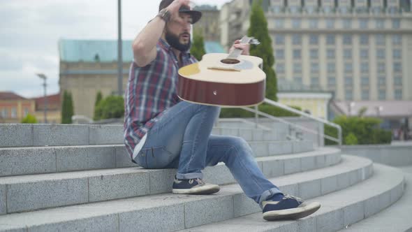 Caucasian Man Walking Up Urban Stairs, Taking Off Hat, and Start Playing Guitar. Portrait of Street alt