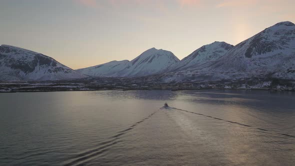 Fishing boat returns to arctic harbour at sunset next to snowy mountains alt