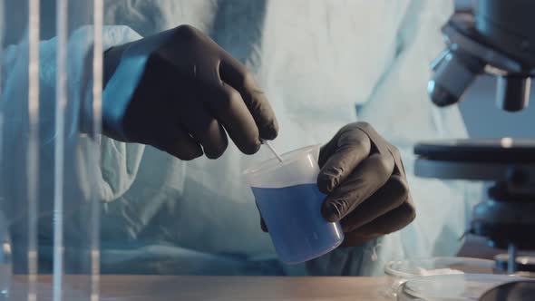 A Laboratory Assistant in Protective Rubber Gloves Stirs a Blue Liquid in a Flask Closeup Against alt