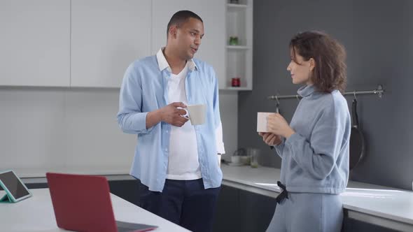 Young Married Couple Standing in Kitchen with Coffee Cups in the Morning Talking alt
