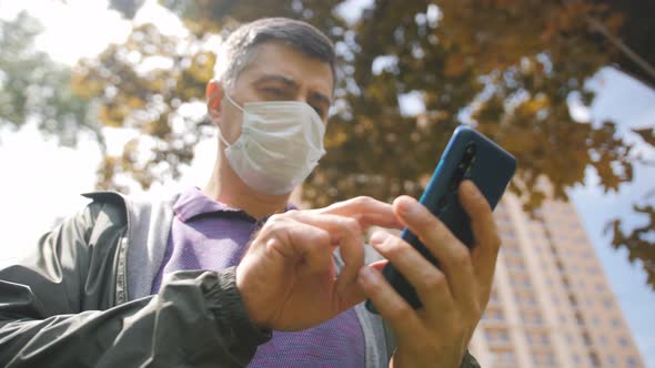 A Middleaged Man in a Medical Protective Mask is Talking on the Phone While Standing on a City alt