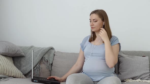 A Pregnant Woman in Home Clothes is Sitting on a Sofa and Using a Laptop alt