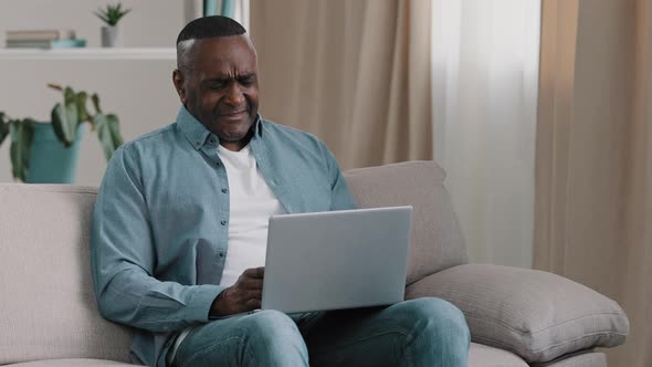 Mature Frustrated African American Man Sitting on Couch in Room Looks at Laptop Screen Reading Bad alt