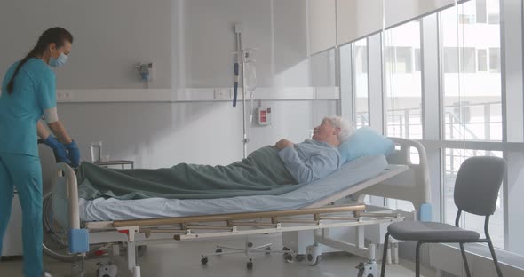 Nurse in Safety Mask Giving Medicine to Elderly Man Patient Lying in Hospital Bed alt