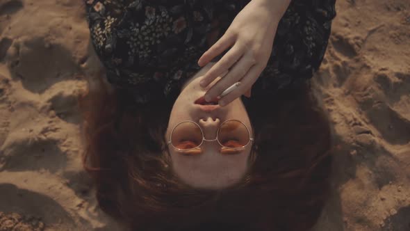 Sad Red-headed Girl Hipster Smokes a Cigarette While Lying on the Sand with His Eyes Closed. Pretty alt