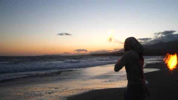 A man in silhouette dancing and spinning a burning fire staff on the beach at sunset with flames and alt