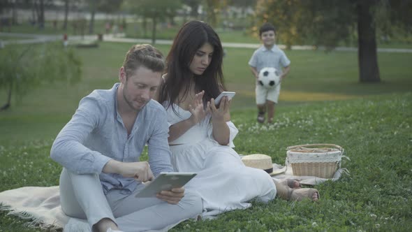 Little Boy Running Ball To Absorbed Parents Sitting Blanket Using Smartphone Tablet alt