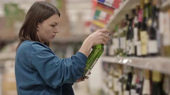 Young Woman Chooses Wine While Standing in Front of Shelves with Alcohol in a Supermarket or Liquor alt
