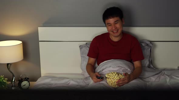 funny young man watching TV and laughing on a bed at night