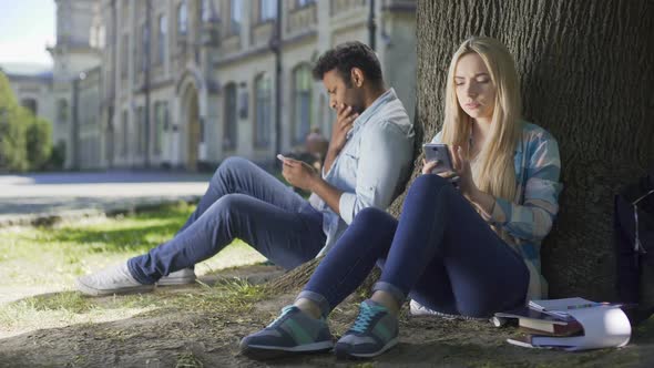 Male, Female Strangers Sitting Under Tree, Using Cellphone, Girl Sighing Heavily alt